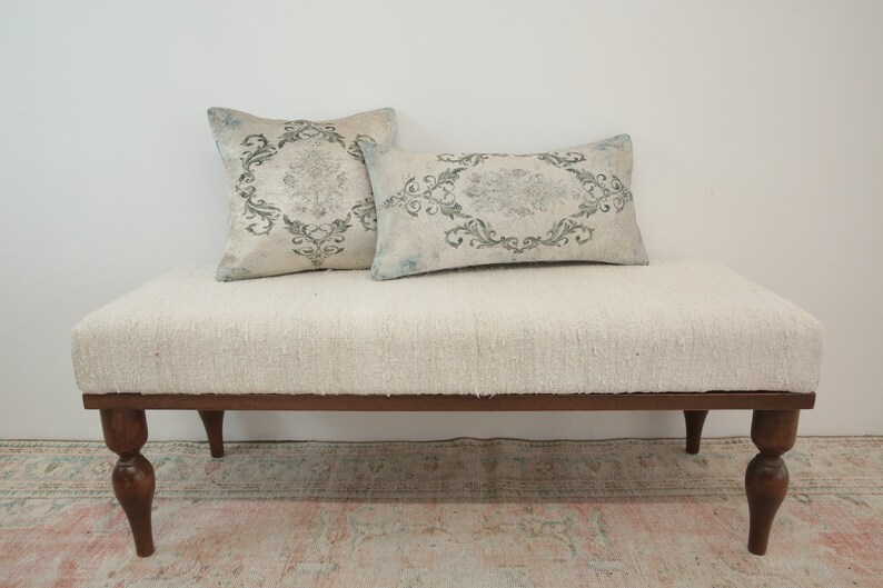 Puede incluir: Un banco tapizado blanco con patas de madera torneadas. El banco tiene dos cojines decorativos con un patr&oacute;n floral en tonos grises y verdes.