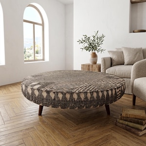 May include: A large, round ottoman with a patterned fabric top in shades of brown and cream. The ottoman has dark wooden legs and is placed in a living room setting. The design features geometric shapes and a decorative edge.