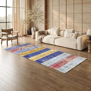 Op de afbeelding: Een rechthoekig vloerkleed met een verweerd, gestreept ontwerp. Het kleed heeft afwisselende banden van blauw, geel, rood en wit, wat een rustieke, verweerde uitstraling geeft. Het ligt op een houten vloer in een woonkamer.