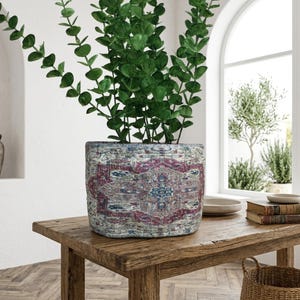 Peut inclure: Un pot de fleurs cylindrique décoratif avec un motif vieilli dans les tons de rouge, bleu et crème. Il contient une plante verte aux longues feuilles fines. Le pot est posé sur une table en bois rustique.