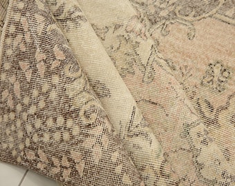 May include: A folded area rug featuring a woven pattern in shades of cream, beige, soft pink, and dark brown. The textile displays intricate floral and geometric motifs across its surface, with a visible texture showing the weave of the fibres.