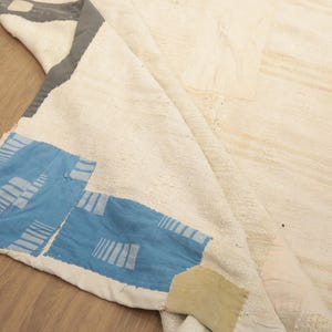 May include: A large off-white area rug with a thick, textured pile. One corner is folded over to reveal a blue fabric patch featuring a white geometric striped pattern, a dark gray border section, and a small tan rectangular fabric accent piece.