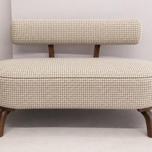 May include: A small loveseat featuring a beige and white houndstooth fabric upholstery. The piece has a rounded, oval-shaped seat and a matching cylindrical backrest supported by two dark brown wooden legs and two wooden back supports.