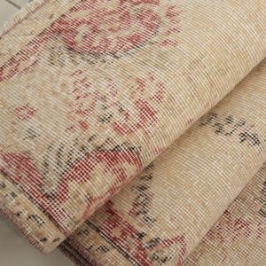 Könnte beinhalten: Nahaufnahme eines beige-roten gemusterten Teppichs mit gewebter Struktur. Der Teppich ist in der Mitte gefaltet, sodass die Unterseite des Stoffes sichtbar ist.