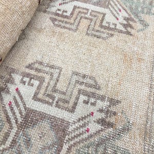 May include: A woven rug featuring a light beige base with repeating geometric patterns in muted brown, gray, and soft green tones. The design includes angular, stepped motifs and small, scattered accents of bright pink thread throughout the weave.