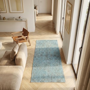May include: A rectangular runner rug featuring a distressed, faded pattern in shades of light blue and grey. The rug is positioned on a light-colored wooden floor with a herringbone pattern, situated in a bright room with neutral-toned furniture.