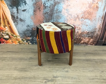 May include: A round, colorful ottoman with a striped fabric cover and wooden legs. The fabric is a mix of red, yellow, green, and blue stripes with a white and brown patterned section.
