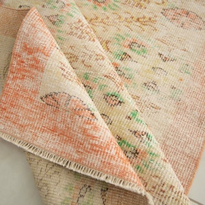 May include: A close-up of a folded rug with a cream and peach color scheme. The rug has a floral pattern with green and brown accents. The rug is made of a woven material and has a fringed edge.