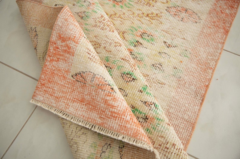 May include: A close-up of a folded rug with a cream and peach color scheme. The rug has a floral pattern with green and brown accents. The rug is made of a woven material and has a fringed edge.