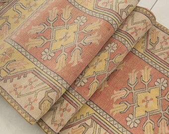 May include: A woven rug featuring a repeating geometric pattern in shades of salmon pink, cream, and pale yellow. The design includes stylized floral motifs and angular borders with thin dark lines, showcasing a textured surface and fringed edges.