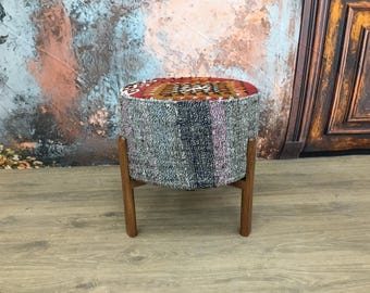 May include: A round, gray and multi-colored patterned ottoman with wooden legs. The ottoman is sitting on a wooden floor.