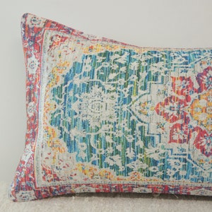 May include: A long, rectangular pillow with a colorful, patterned design. The pattern features a large, central floral motif in shades of red, yellow, and blue, surrounded by smaller floral and geometric designs. The pillow is made of a soft, textured fabric.
