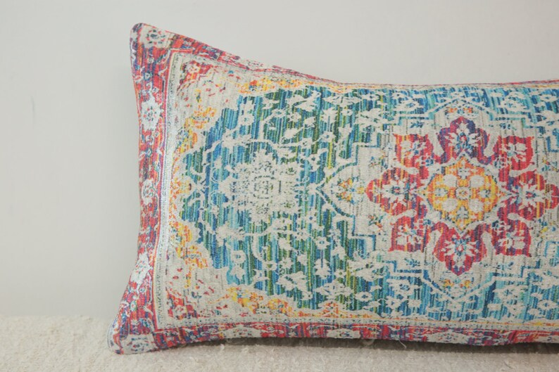 May include: A long, rectangular pillow with a colorful, patterned design. The pattern features a large, central floral motif in shades of red, yellow, and blue, surrounded by smaller floral and geometric designs. The pillow is made of a soft, textured fabric.