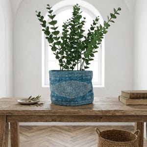 May include: A blue fabric planter with a light blue oval design holds a lush green plant. The planter sits on a rustic wooden table, with a small plate and books nearby. The background features a large arched window.