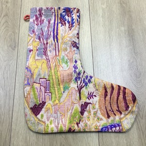 Può includere: Un calzino di Natale colorato con un design intrecciato che presenta una varietà di animali, piante e motivi geometrici. Il calzino è realizzato in un tessuto morbido e testurizzato e ha uno sfondo bianco con accenti di viola, rosa, giallo, verde e blu.