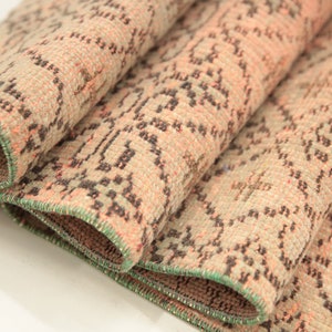 May include: Close-up of a folded rug with a beige background and a repeating pattern of small black and pink shapes. The rug has a green border.