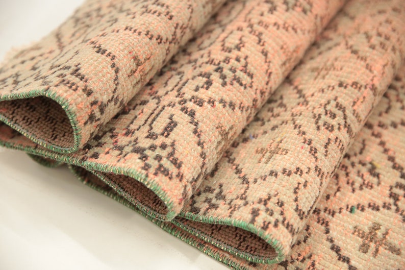 May include: Close-up of a folded rug with a beige background and a repeating pattern of small black and pink shapes. The rug has a green border.