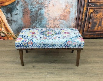 May include: A rectangular ottoman featuring a vibrant, multi-colored patterned fabric top with shades of blue, pink, and yellow. The piece is supported by four tapered, dark brown wooden legs, providing a stable base for the upholstered seat.
