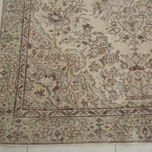 K&ouml;nnte beinhalten: Ein Vintage-Teppich mit beigefarbenem Hintergrund und einem floralen Muster in Gr&uuml;n und Braun. Der Teppich hat einen verblassten Look und eine abgenutzte Textur.