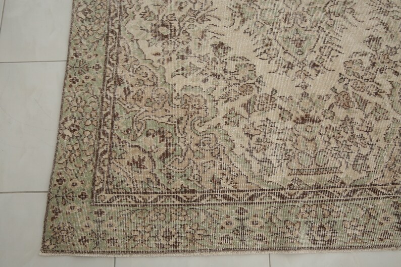 K&ouml;nnte beinhalten: Ein Vintage-Teppich mit beigefarbenem Hintergrund und einem floralen Muster in Gr&uuml;n und Braun. Der Teppich hat einen verblassten Look und eine abgenutzte Textur.