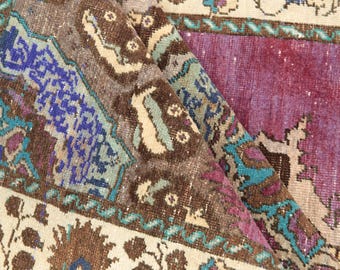 May include: A woven rug featuring a complex pattern with deep purple, brown, cream, and bright turquoise blue sections. The design includes geometric shapes and intricate floral-inspired motifs arranged in a layered, folded composition.