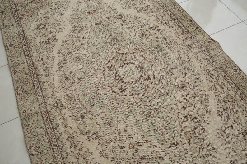 K&ouml;nnte beinhalten: Ein Vintage-Teppich mit beigefarbenem Hintergrund und einem braunen und gr&uuml;nen floralen Muster. Der Teppich hat ein zentrales Medaillon mit einem Sternmuster.