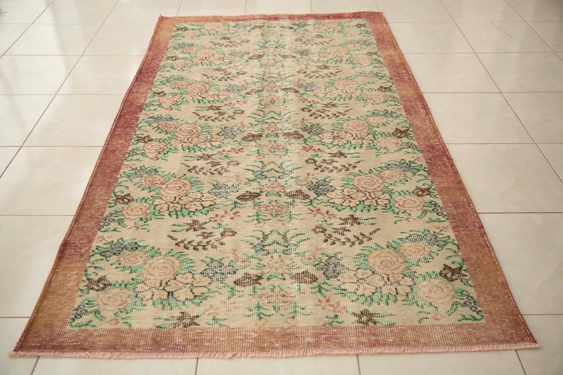 May include: A vintage floral rug with a cream background and a pink border. The rug features a repeating pattern of pink, green, and brown flowers.