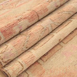 May include: A folded area rug featuring a textured pile with a faded pattern in shades of soft peach, cream, and muted brown. The rug displays a repeating geometric and floral motif across its surface, with visible fiber density and weave.