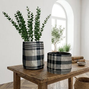 Pode incluir: Duas floreiras de tecido com um padrão xadrez em preto, cinza e bege. A floreira maior contém caules verdes e folhosos. As floreiras estão sobre uma mesa de madeira. A floreira menor está vazia.