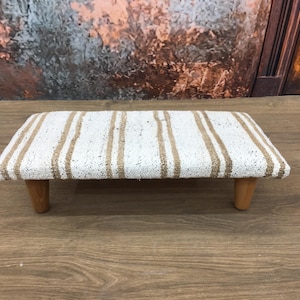 May include: A small, rectangular footstool with a white and brown striped fabric top and wooden legs.