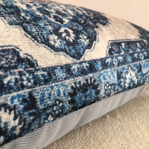 Op de afbeelding: Decoratief kussen met een blauw en wit patroon. Het kussen heeft een gedetailleerd, sierlijk patroon met tinten blauw en wit. Het kussen heeft een grijze stoffen rand.