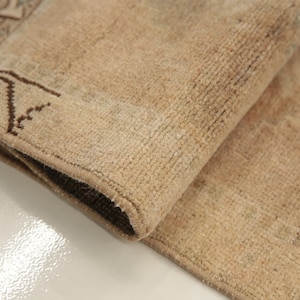 May include: Close-up of a beige rug with a brown patterned border. The rug is folded in half, showing the texture of the fabric.