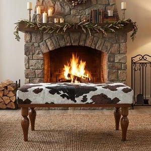 May include: A bench with a cowhide pattern top and wooden legs is positioned in front of a stone fireplace with a burning fire. The bench has a brown and white pattern. The fireplace is decorated with candles and greenery.