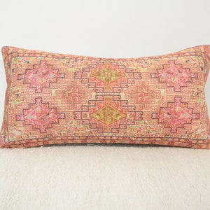 May include: A long rectangular pillow with a pink, red, and yellow geometric pattern. The pillow has a cream-colored background and a dark brown border.