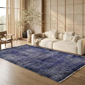 Pode incluir: Um tapete azul desgastado, retangular e grande, com uma aparência vintage desbotada. O tapete está colocado sobre um chão de madeira numa sala de estar, com um sofá branco, uma cadeira de madeira e paredes e cortinas em tons neutros.