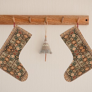 May include: Two green and brown patterned Christmas stockings hang from a wooden rack with a small, gray, fabric Christmas tree ornament hanging in the middle.