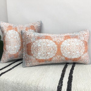 May include: Two decorative throw pillows with a white and orange patterned design. The pillows are on a white and black striped surface.