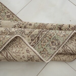 K&ouml;nnte beinhalten: Ein gefalteter Vintage-Teppich mit beigefarbenem Hintergrund und einem braunen und gr&uuml;nen Blumenmuster.