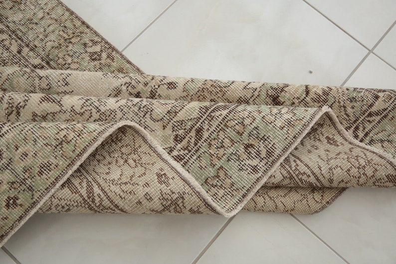 K&ouml;nnte beinhalten: Ein gefalteter Vintage-Teppich mit beigefarbenem Hintergrund und einem braunen und gr&uuml;nen Blumenmuster.