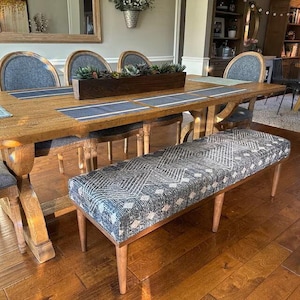 May include: A rectangular upholstered bench featuring a dark blue and white geometric diamond pattern fabric, supported by four tapered wooden legs. The bench is positioned in front of a large wooden dining table with matching chairs.