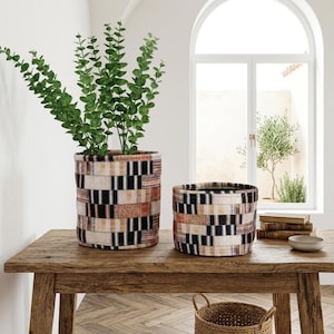 Könnte beinhalten: Zwei zylindrische Stofftöpfe mit einem Patchwork-Design aus schwarzen, weißen und beigefarbenen Rechtecken. Ein Topf enthält eine hohe grüne Pflanze. Die Töpfe stehen auf einem rustikalen Holztisch, im Vordergrund ein Weidenkorb.