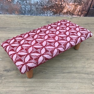 Peut inclure: Un petit repose-pieds rectangulaire avec un motif floral rouge et blanc sur le coussin. Le coussin est soutenu par quatre pieds courts en bois. Le repose-pieds est posé sur une surface en bois.