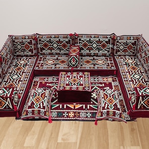 Puede incluir: Un juego de asientos de suelo burdeos con intrincados patrones geométricos en blanco, naranja y verde. El conjunto incluye múltiples cojines, respaldos y una alfombra central, todos adornados con borlas. El diseño recuerda la decoración tradicional de Oriente Medio.