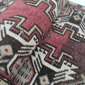 May include: A woven rug featuring a geometric pattern in shades of deep red, dark brown, and cream. The textile displays a repeating angular motif with sharp lines and symmetrical shapes, showing a folded corner that reveals the textured pile surface.
