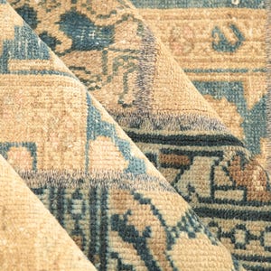 May include: A folded textile rug featuring a repeating geometric pattern in shades of muted blue, tan, and beige. The fabric displays a dense, woven texture with visible pile and intricate linear motifs across the surface of the folded material.