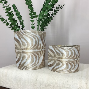 Puede incluir: Dos contenedores de almacenamiento cilíndricos de tela con un patrón de cebra en tonos marrones y blancos. Los contenedores están sobre una superficie blanca.