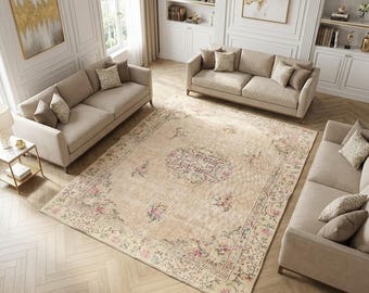 Puede incluir: Una alfombra rectangular grande con base beige, un medallón central y motivos florales en tonos rosa, verde y marrón. La alfombra está situada en el centro de una habitación con tres sofás de color beige y suelo de madera clara.