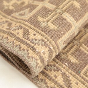 May include: A woven rug featuring a taupe and beige geometric pattern with a repeating border design. The textile has a thick, textured pile with visible knotted construction and a folded corner showing the underside and finished edge.