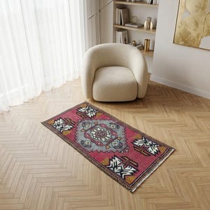 Può includere: Un tappeto rettangolare in stile vintage con un motivo a medaglione centrale. Il tappeto presenta un campo prevalentemente rosso con motivi geometrici bianchi, neri e oro. È posizionato su un pavimento in legno chiaro, vicino a una poltrona color crema.