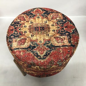 May include: A round ottoman with a colourful, patterned fabric top. The fabric features a red, blue and yellow design with intricate details. The ottoman has a wooden base.